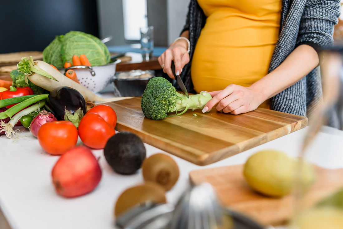 A pregnant woman is standing in the kitchen cutting fruits and vegetables on a wooden board