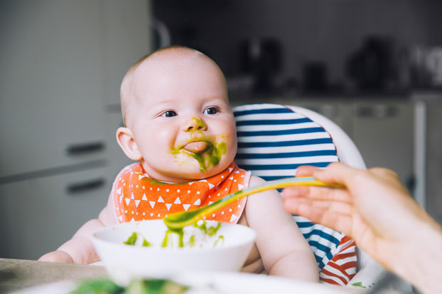 Baby sitting in high chair is fed with green puree